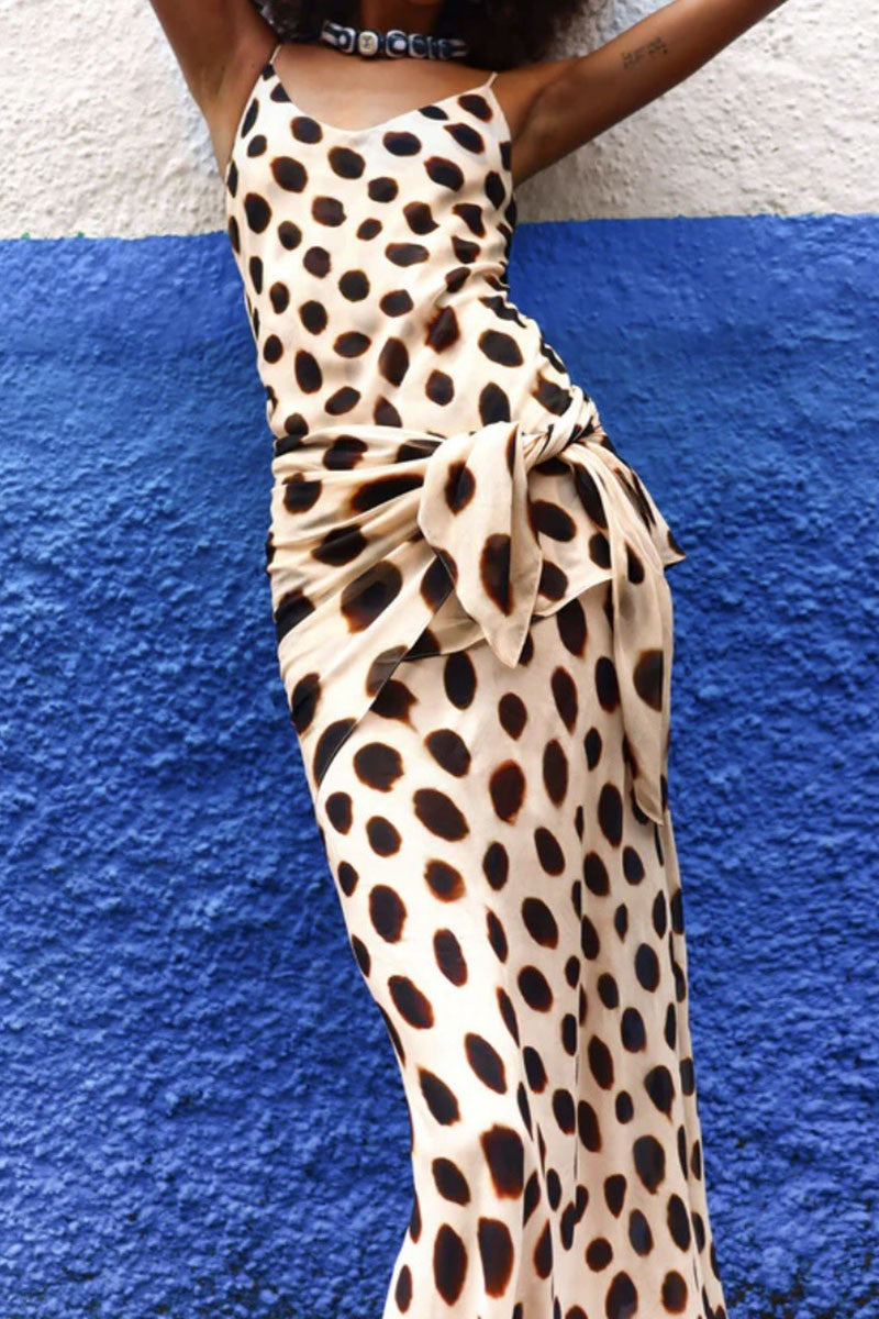 A sleeveless beige maxi dress with a bold black leopard print pattern, featuring a side tie detail, worn by a model standing against a textured blue and white wall.