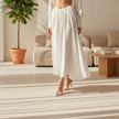 A model wearing a flowing white high-waisted midi skirt with a smocked waistband, paired with white heeled sandals, set against a textured gray studio background.