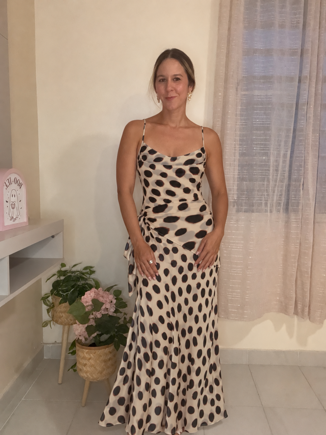 A woman modeling a sleeveless maxi dress with a beige base and black leopard print spots, standing in an indoor setting with a window and sheer curtains behind her.