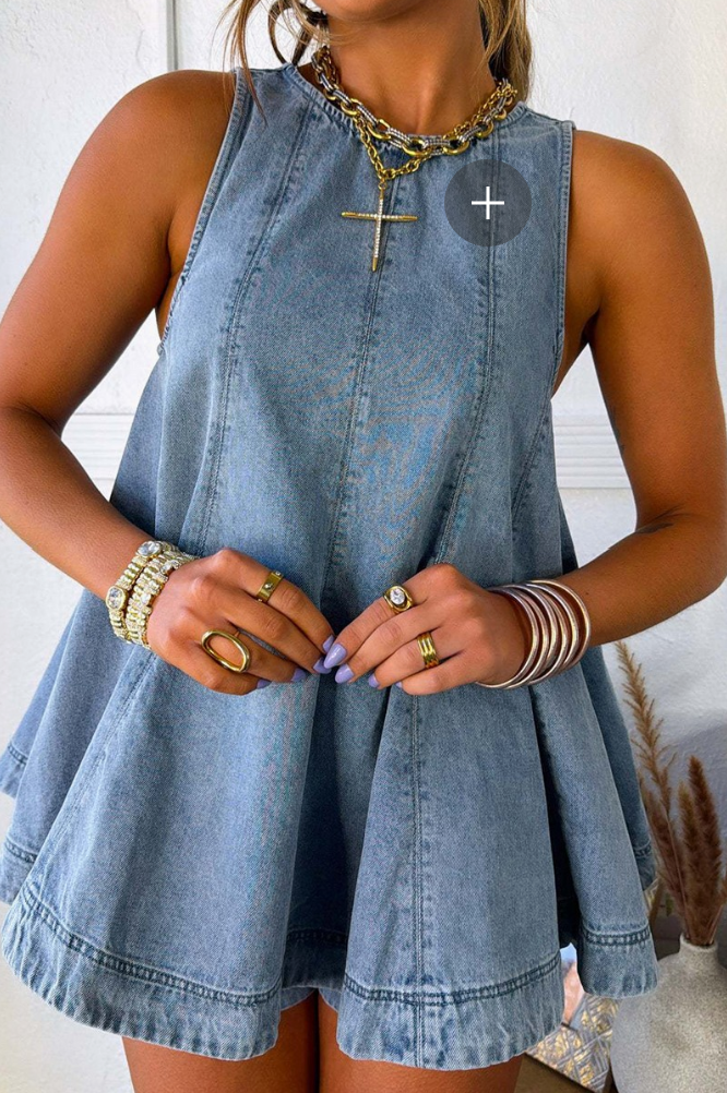 A woman wearing a sleeveless light blue denim mini dress with a flared peplum skirt, styled with layered gold necklaces and bracelets.