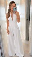 A woman takes a mirror selfie wearing a white sleeveless wide-leg jumpsuit with a deep V-neckline in a brightly lit hallway.