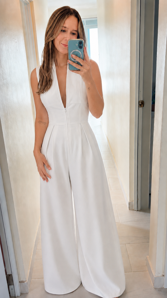 A woman takes a mirror selfie wearing a white sleeveless wide-leg jumpsuit with a deep V-neckline in a brightly lit hallway.