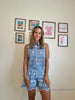 Woman wearing a blue floral sleeveless top and shorts standing in front of a wall with framed pictures.