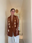 A woman wearing a sleeveless rust-brown blazer vest with decorative tassel details down the front, paired with white pants, standing in a hallway.