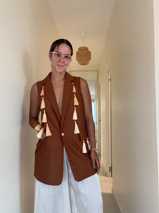 A woman wearing a sleeveless brown blazer vest with beige tassel details and white pants, standing in a hallway.