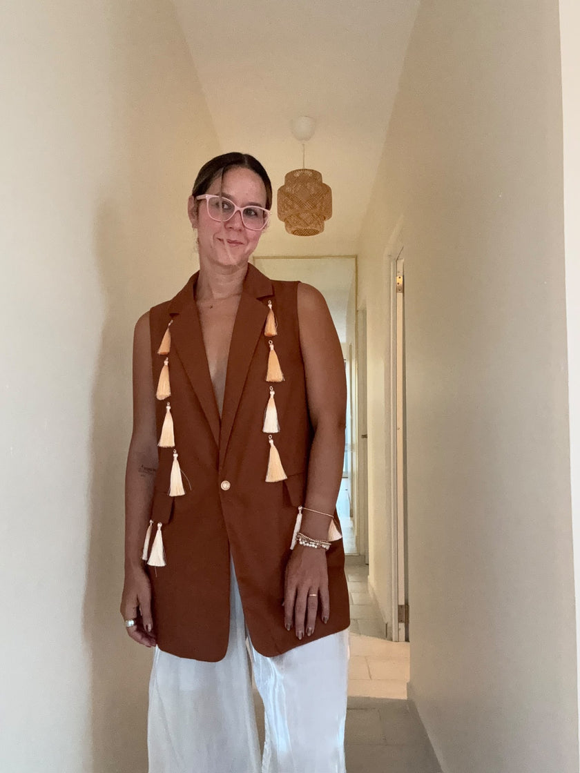 A woman wearing a sleeveless rust-brown blazer vest with decorative tassel details down the front, paired with white pants, standing in a hallway.
